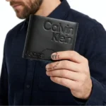 Man in a dark shirt holding a black leather Calvin Klein wallet with embossed logo visible.