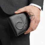 Man in a dark suit holds a black leather wallet with a large embossed CK logo visible on the front edge.