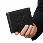 Black leather wallet with embossed CK logo held by a hand against a white background.
