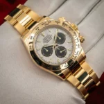 Gold Rolex Daytona chronograph wristwatch with a tachymeter bezel and three black subdials on a silver-toned face, resting on light fabric surface.