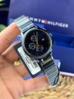 Tommy Hilfiger wristwatch with black dial, gold hands, and metal band held in hand against a blue box.