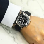 Wrist wearing a black-dial Rolex Daytona with diamond markers and a black rubber strap, resting on a marble surface while a white cuff shows on the sleeve.