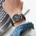 Silver and black chronograph watch on a wrist with a black rubber strap and denim cuff visible in the foreground, studio shot