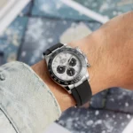 Wrist wearing a silver Rolex Daytona chronograph with a black bezel and black rubber strap, denim sleeve visible.