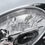 Close-up of a Rolex watch dial featuring the crown logo and engraved text, with a textured silver surface and subdials nearby.