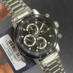 1792181-tommy-hilfiger-watch-men-black-dial-metal-stainless-steel-silver-strap-quartz-analog-rocky_10 Silver metal wristwatch with a black dial, three subdials, and a Tommy Hilfiger logo on the face, held against a dark card label with a barcode.