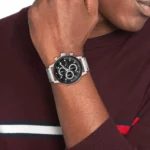 1792181-tommy-hilfiger-watch-men-black-dial-metal-stainless-steel-silver-strap-quartz-analog-rocky_4 Silver stainless steel chronograph watch on a wrist, black dial with subdials, worn with a maroon sweater outfit.
