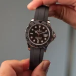 Close-up of a Rolex Yacht-Master watch with a black dial and black rubber strap, held by two hands; date window at 3 o'clock magnified by the cyclops lens