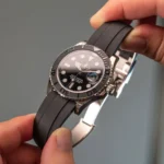 Close-up of a Rolex Yacht-Master watch with a black dial, ceramic bezel, and black rubber strap held by fingers.
