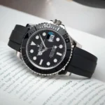 Close-up of a Rolex Yacht-Master wristwatch with a black dial, white markers, and a black rubber strap on a printed page, showing the date window.