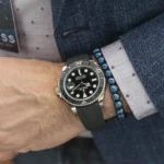 Wrist close-up with a black-dial Rolex diver's watch, gray bezel, and rubber strap, paired with a blue beaded bracelet under a navy blazer and light purple polka-dotted shirt cuff.