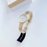 Gold wristwatch with a metallic link band and white face, placed on a white surface with a box nearby in the background.