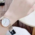 Gold-toned wristwatch with a white dial and crystal hour markers on a woman's wrist, with a mesh band.