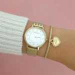 Gold-tone wristwatch with a white dial, paired with a gold link bracelet and a heart 'Mom' charm bracelet against a pink background.