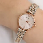 Two-tone rose gold and silver wristwatch on a wrist, white dial with Roman numerals and crystal markers, Emporio Armani logo visible.