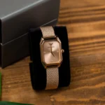 ar11347-emporio-armani-watch-square-women-rose-gold-dial-mesh-stainless-steel-metal-rose-gold-strap-quartz-battery-analog-gioia_20 Rose-gold rectangular Emporio Armani wristwatch with diamond markers on a black display cushion, resting on a wooden surface.