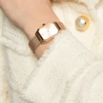 ar11347-emporio-armani-watch-square-women-rose-gold-dial-mesh-stainless-steel-metal-rose-gold-strap-quartz-battery-analog-gioia_400 Rose gold square-faced wristwatch with a mesh band on a wrist, resting on a fluffy white sweater.