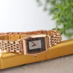 Rose-gold rectangular wristwatch with a diamond-studded center, resting on a gold surface.