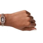 Gold-toned bracelet wristwatch on a left wrist with a rectangular black dial and diamond-studded center.