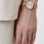 Rose gold wristwatch with a diamond-studded face on a person's left wrist, beige sleeve in background.