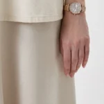 Gold-toned wristwatch on a person’s left wrist, paired with a beige top and skirt, with the hand resting at the side.