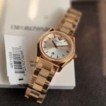 Rose gold-tone women's wristwatch with a diamond-studded bezel resting on a white box, displaying the Emporio Armani logo on the dial