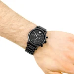 Black metal chronograph wristwatch worn on a forearm with visible hairs and a metal link band