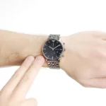 Close-up of a silver metal wristwatch with a black dial and chronograph subdials on a wrist, as another hand touches the wrist.