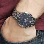 Silver stainless-steel chronograph wristwatch on a man’s left wrist, burgundy shirt and denim jeans visible in the background.