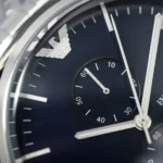 Close-up of a dark blue luxury wristwatch dial featuring a silver Emporio Armani eagle logo, white hour markers, and silver hands.