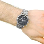 Wrist wearing a silver metal chronograph watch with a black dial and Emporio Armani logo, on a light forearm against a white background