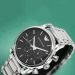Close-up of a silver-toned Emporio Armani chronograph watch with a black dial and metal bracelet against a green background.