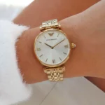 Gold-toned wristwatch with a white face and Roman numerals on a metal link bracelet worn on the wrist (Emporio Armani logo visible).