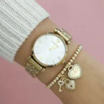 Gold wristwatch with a white dial and Roman numeral markers, worn with multiple gold bracelets on a pink background.