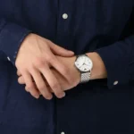 Person in a dark blue shirt resting their hands, showing a silver metal wristwatch with a white face and Roman numerals on the left wrist.