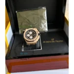 Gold-tone Audemars Piguet watch with black dial, displayed in an open black presentation box beside a wrapped item and booklet behind it.