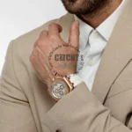 Man in a beige suit adjusts a rose-gold chronograph watch on his left wrist, white shirt visible underneath.