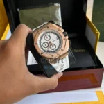 Rose gold chronograph wristwatch with a white patterned dial and three subdials, held in a hand over a black presentation box.