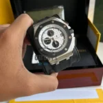Hand holding a luxury chronograph watch with a white textured dial, black bezel, and metal case, in front of a box.