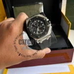 audemars-piguet-watch-battery-diver-for-men-silver-metal-case_2 Close-up of a luxury chronograph watch on a wrist, stainless steel case, black dial with multiple subdials, and a black strap, with a box in the background.