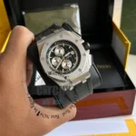 audemars-piguet-watch-battery-for-men-silver-metal-case-silver-chronograph_2 Close-up of a stainless steel chronograph wristwatch with a black dial and rubber strap being held, with a watch box in the background.