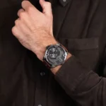 Close-up of a man wearing a dark shirt and a large gray chronograph watch with a brown leather strap on his wrist.