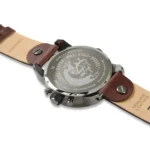 Back of a stainless steel watch with a brown leather strap, showing the engraved case back and side crown/buttons on a white background.