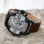 Large men's chronograph watch with gray dial, black bezel, and brown leather strap on a rocky surface.