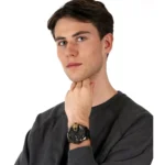 dz7377-original-diesel-watch-balck-leather-strap_4 Portrait of a young man in a dark sweater, touching his chin with his left hand, wearing a black wristwatch with gold accents on his wrist.