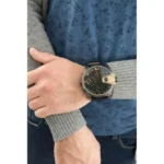 dz7377-original-diesel-watch-balck-leather-strap_6 Close-up of a man's wrist with a large black watch featuring gold 12 and 6 numerals, worn over a gray knit sleeve with a blue patterned shirt in the background.