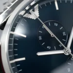 Close-up of a blue watch dial with silver hands and a small circular subdial showing seconds or minutes, and a silver bezel edge visible on the left.