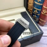 Close-up of a Hublot watch clasp on a black rubber strap, held by a hand with a blurred bookcase in the background.