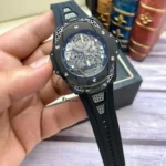 Skeleton wristwatch with exposed gears and a black case held in a hand; black rubber strap with gray vented accents.
