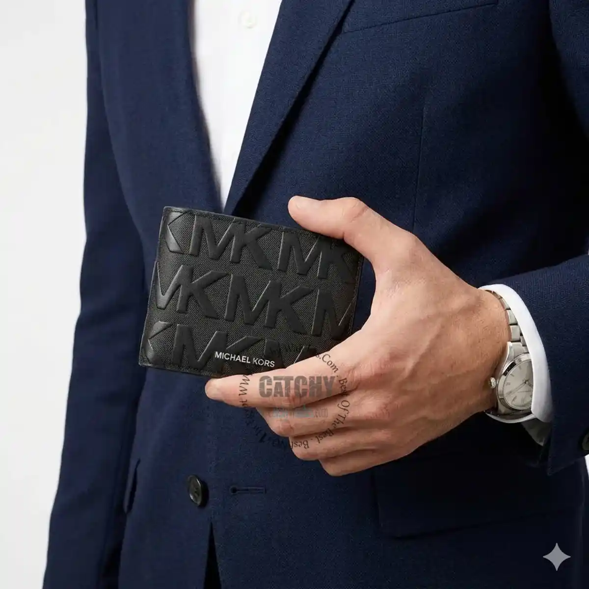Close-up of a man in a navy suit holding a black Michael Kors wallet with the MK pattern visible on the surface.